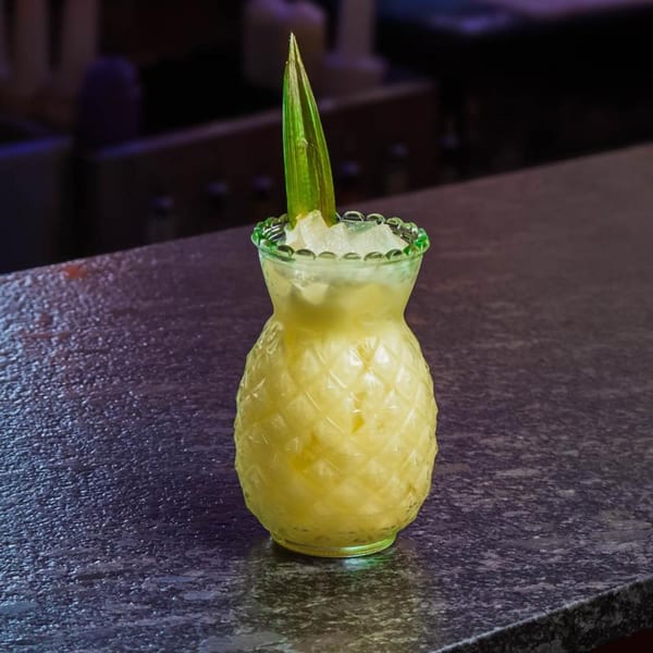 A pineapple-shaped glass filled with a yellow drink, ice, and a green leaf garnish on a dark counter.