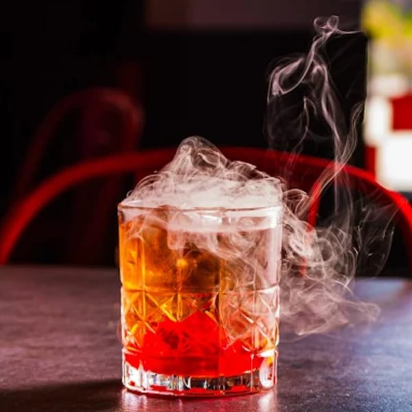 A smoking cocktail with a red cherry at the bottom, on a dark table with red accents.