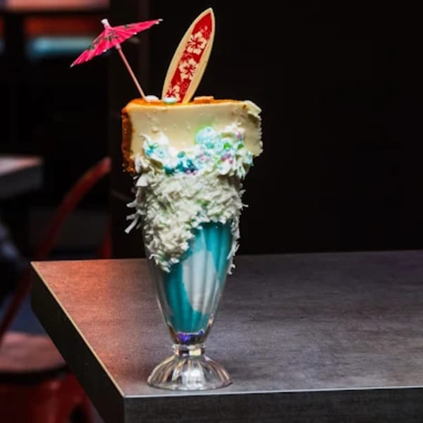 A cheesecake slice on a blue milkshake, topped with a surfboard and pink umbrella.